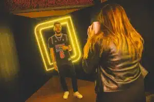 A man poses in front of a neon yellow frame while a woman takes his photo; he wears a basketball jersey and sneakers.