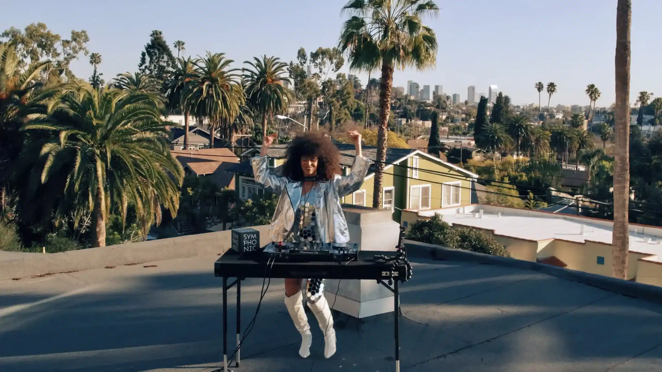 A person with curly hair performs as a DJ on a rooftop with palm trees, houses, and a city skyline visible in the background.