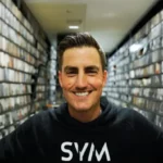 A man in a black SYM hoodie smiles at the camera while standing in a narrow hallway lined with shelves full of organized items.