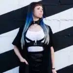 Woman with long ombre blue hair and bangs, wearing a white top, black harness, and black skirt, stands against a white and black diagonal striped wall.